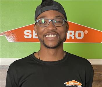 Male employee with a hat and glasses smiling in front of green SERVPRO background.